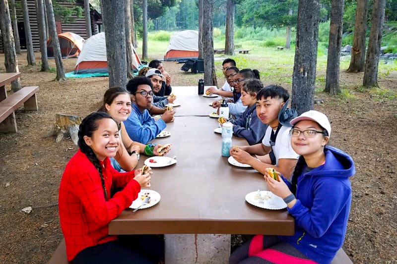 high school students eating a meal on picnic tables at a campsite