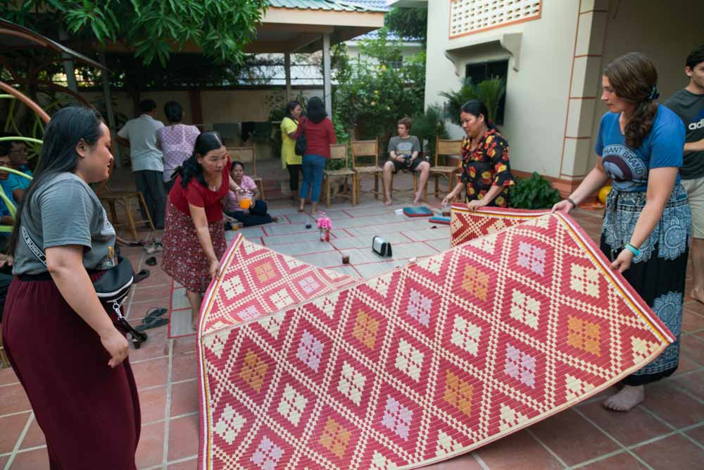 Women in Cambodia spread out colorful mat in courtyard.