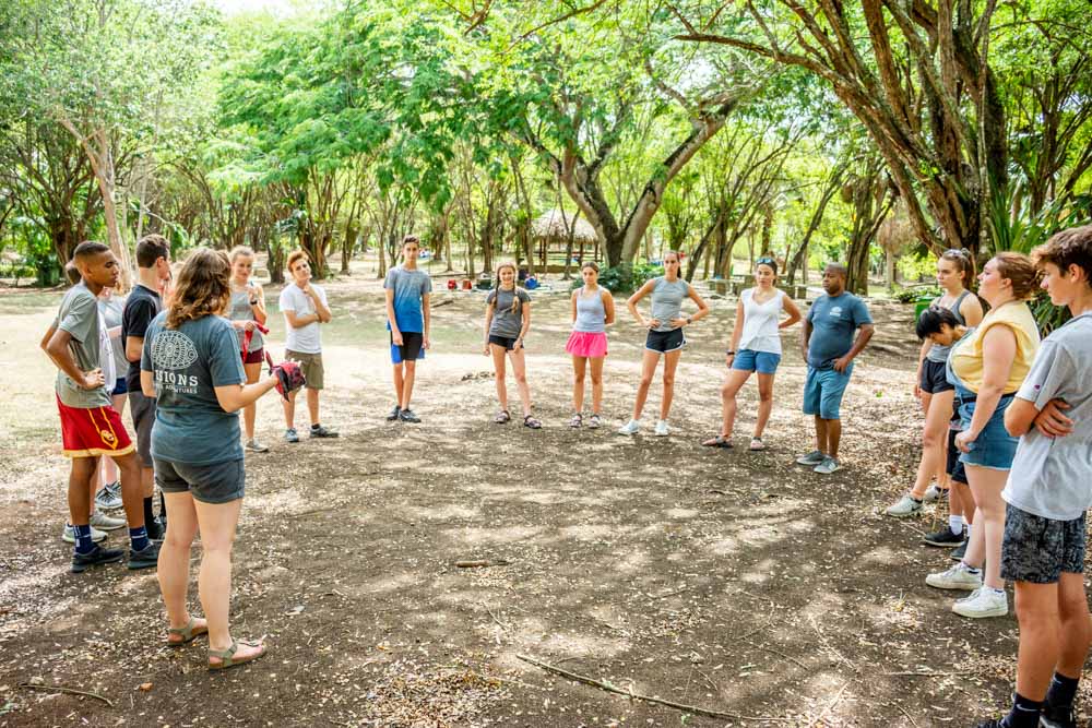 A group of teen volunteers stands in a circle in the forest to discuss service projects.