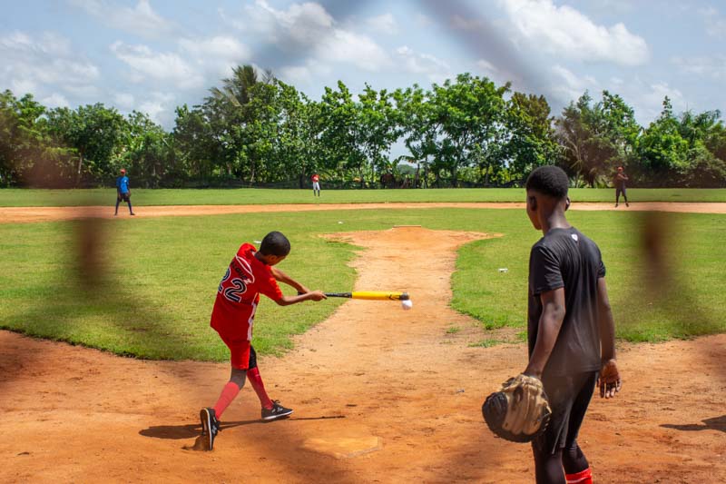 Looking through chain link fence, boy hits baseball in the Dominican Republic