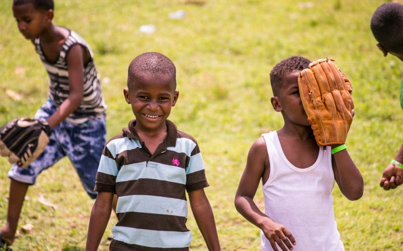Dominican kids smile on baseball diamond as one partially covers his face with a baseball glove
