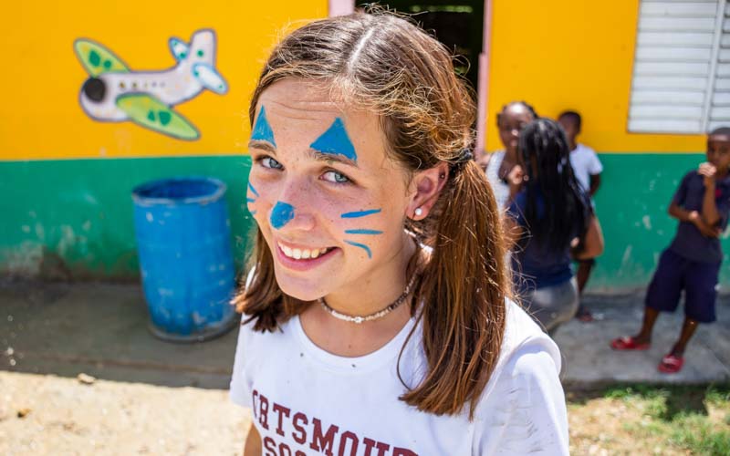 Teen volunteer with cat-like face paint smiles with children in the background