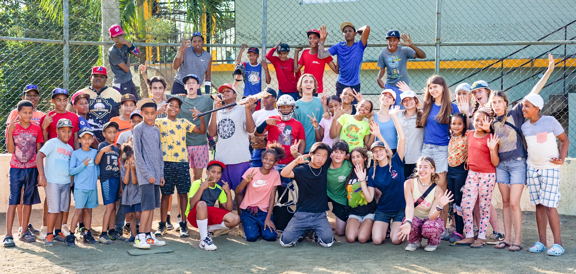 A large, diverse group of children and teenagers pose together on a baseball field, smiling and making playful gestures in front of a chain-link fence on a sunny day. Some wear baseball caps and sports gear.