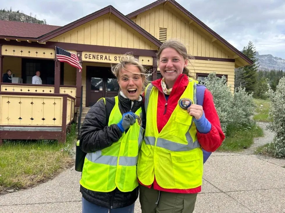 Two smiling women wearing yellow safety vests stand outside a rustic building labeled General Store. One holds a badge, and both appear happy. An American flag hangs from the porch, and trees and mountains are in the background.
