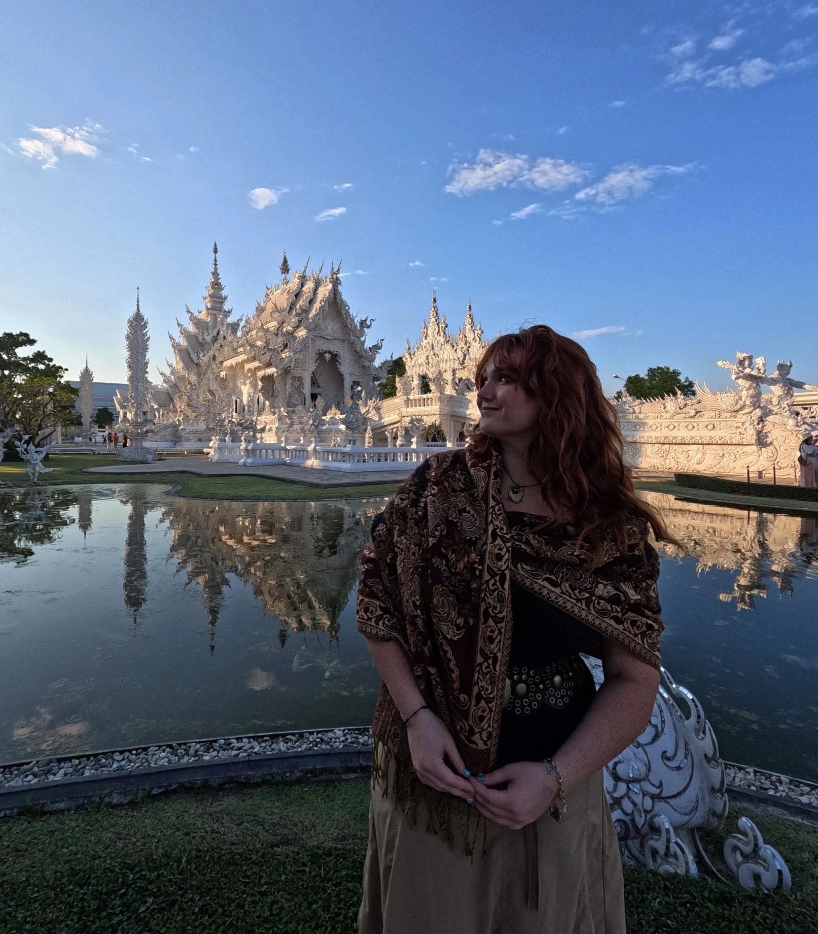 A woman stands by a reflective pond in front of the ornate, white Wat Rong Khun temple in Thailand, under a blue sky with scattered clouds. She wears a patterned shawl and looks to her left, smiling.