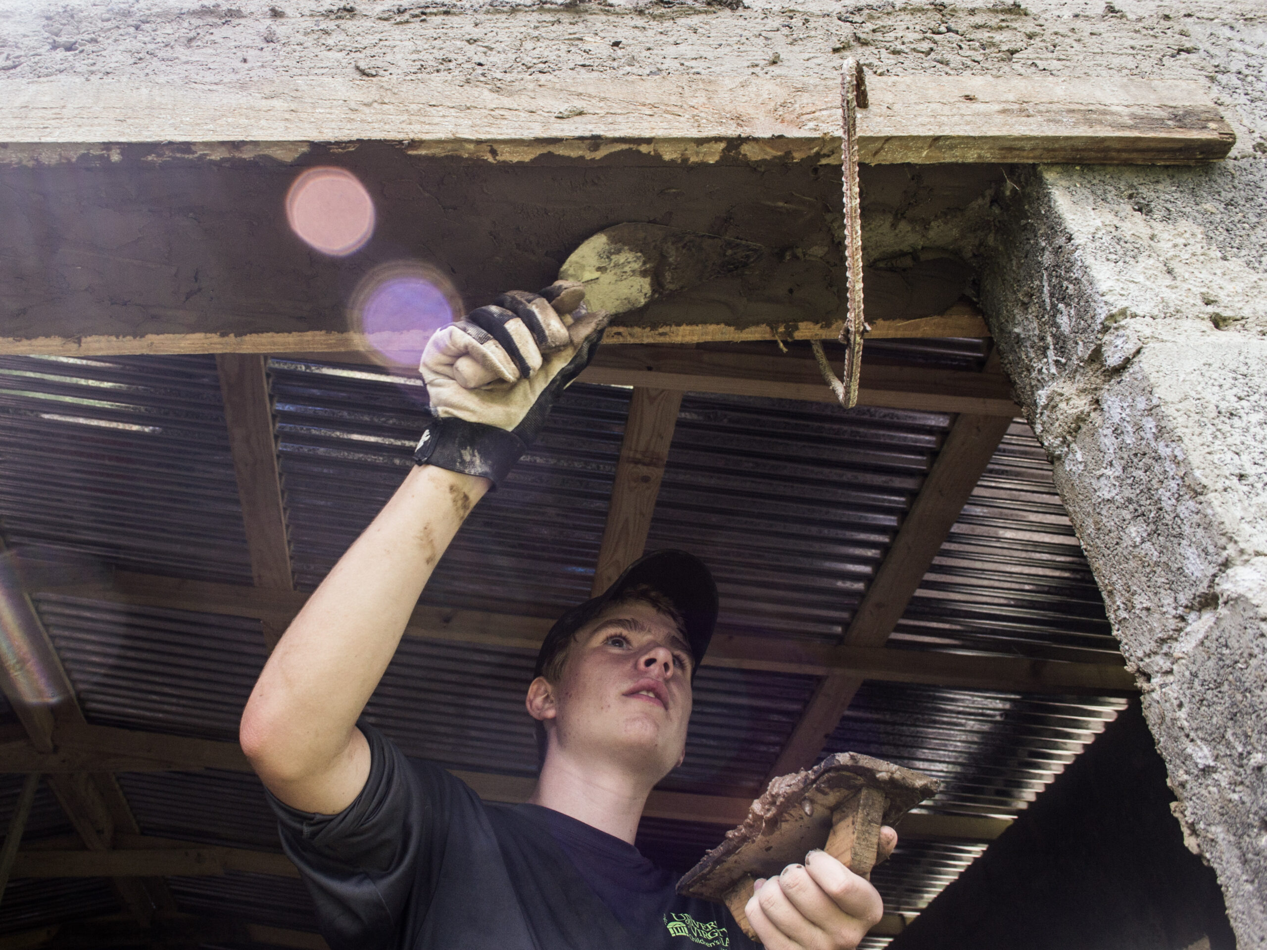 A teenager applying mud or plaster to the top of a doorway while helping construct a building with a corrugated metal roof - VISIONS Summer programs
