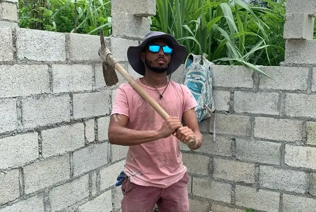A man wearing sunglasses, a wide-brim hat, and a pink shirt holds a pickaxe while standing inside a partially built brick structure, with tall green plants in the background. A backpack hangs on the wall behind him.