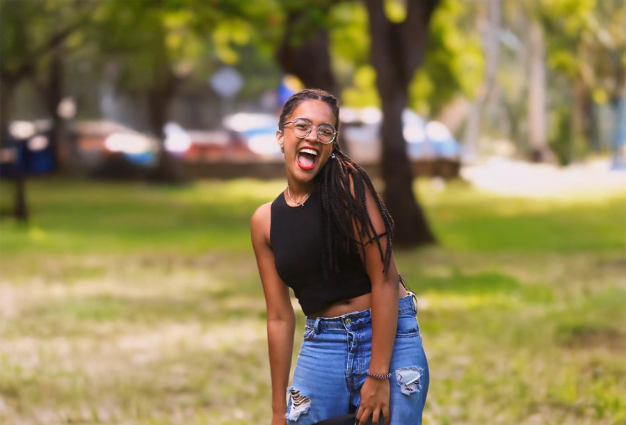 A young woman with braids, glasses, and a black sleeveless top stands outdoors on grass, smiling widely with her tongue out. She is wearing ripped blue jeans and appears happy, with trees and a blurred background behind her.