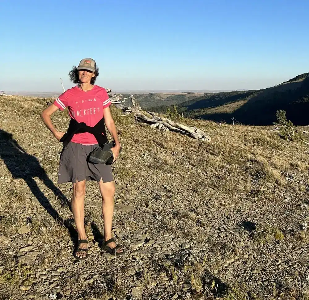 A person wearing a red t-shirt, shorts, sandals, and a cap stands on a rocky hillside in the sunlight, with shadows cast on the ground and rolling hills in the background.