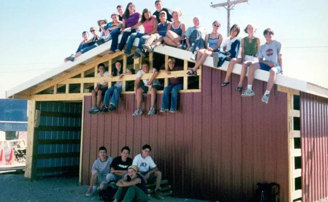 A group of people sit on the roof and in front of a red and tan wooden shed, posing and smiling for a photo on a sunny day. The sky is clear and power lines are visible in the background.