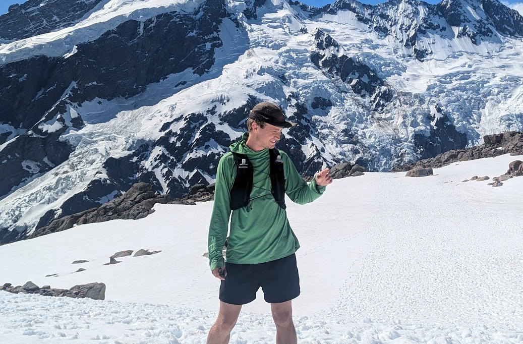 A person wearing a green long-sleeve shirt, black shorts, and a cap stands on snowy terrain with tall, rugged snow-covered mountains in the background.