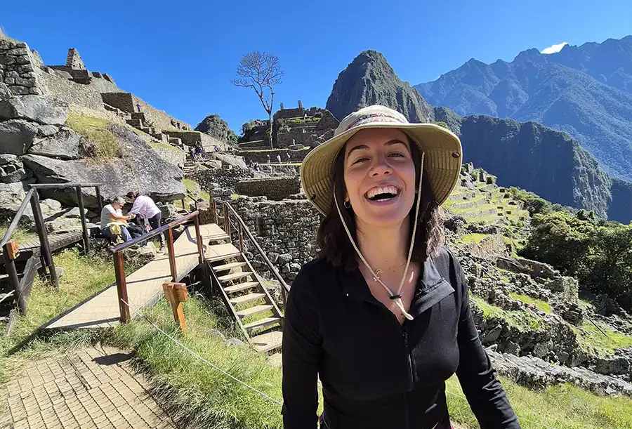 A smiling woman in a sunhat stands at the ruins of Machu Picchu in Peru, with stone terraces, mountains, and a clear blue sky in the background. Other visitors are exploring the ancient site nearby.