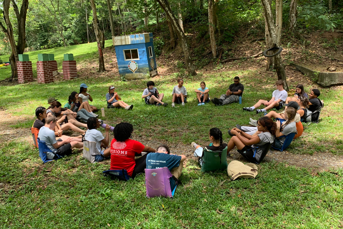 A group of people, mostly children and a few adults, sit in a large circle on grass in a wooded area, participating in an outdoor group activity. A small blue building and trees are visible in the background.