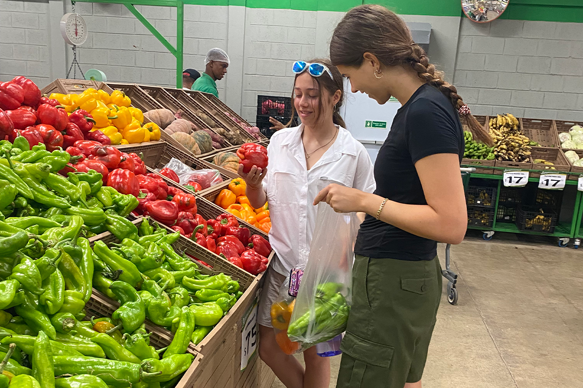 Two young women stand by a display of peppers at a grocery store, selecting green and red peppers, while a man shops in the background. One holds a plastic bag with peppers inside.