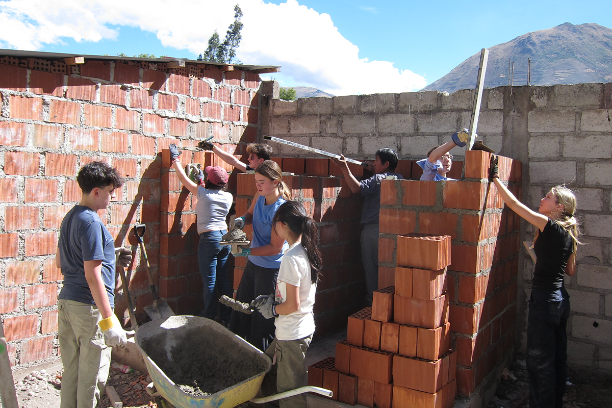 A group of people work together building a brick wall outdoors on a sunny day, using tools and wearing gloves. A wheelbarrow with cement is in the foreground, with mountains and blue sky in the background.