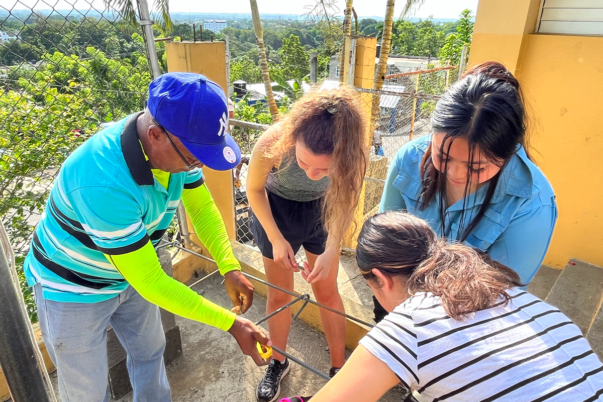 Four people work together outdoors, focused on assembling or tying something with black cords or ropes, next to a yellow building and metal fence, with greenery and a distant landscape in the background.
