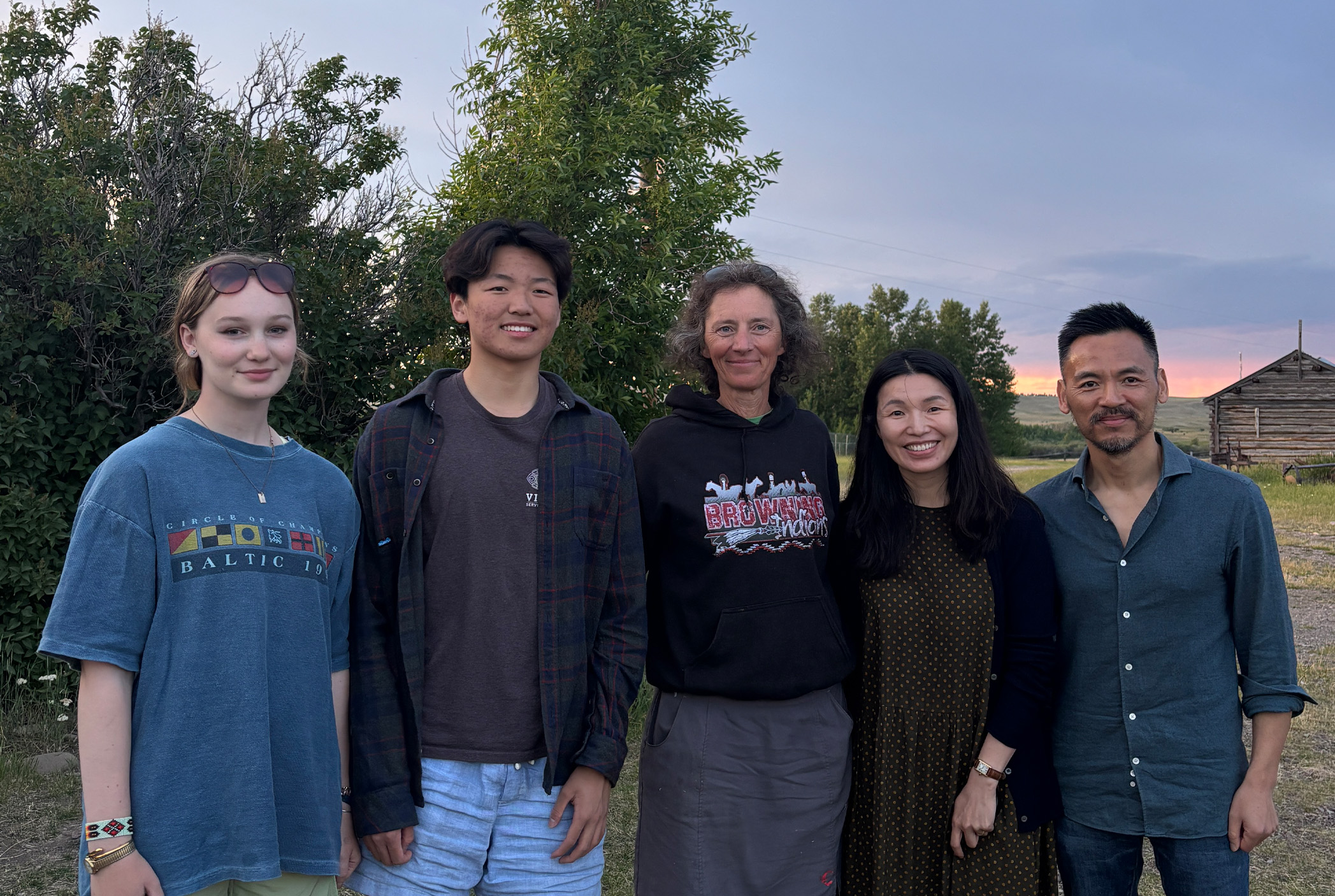 Five people stand outside in a row, smiling at the camera. They are dressed casually and are standing in front of trees and a rustic wooden building at sunset.