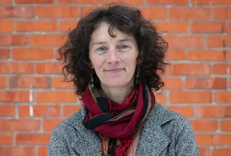 A woman with curly brown hair stands in front of a red brick wall, wearing a gray coat and a colorful scarf. She is looking at the camera and smiling softly.