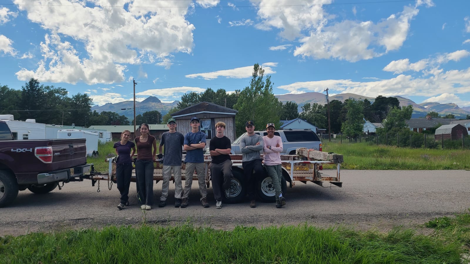 Six people stand in a row in front of a trailer loaded with materials, which is attached to a pickup truck. They are outdoors on a sunny day with mountains, trees, and houses in the background.
