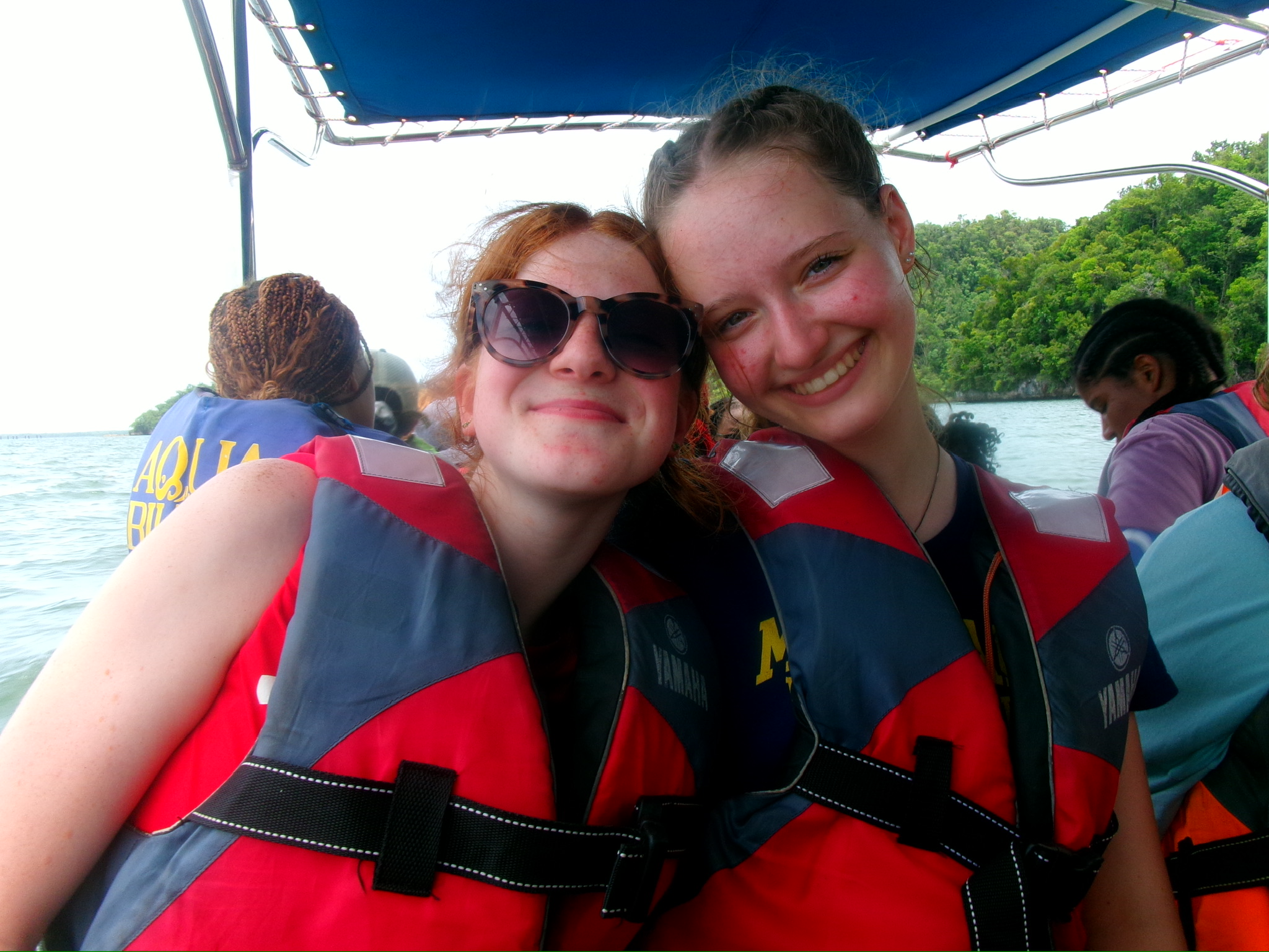 Two smiling young people wearing red and blue life jackets are sitting close together on a boat. One wears sunglasses. Other passengers and greenery are visible in the background.