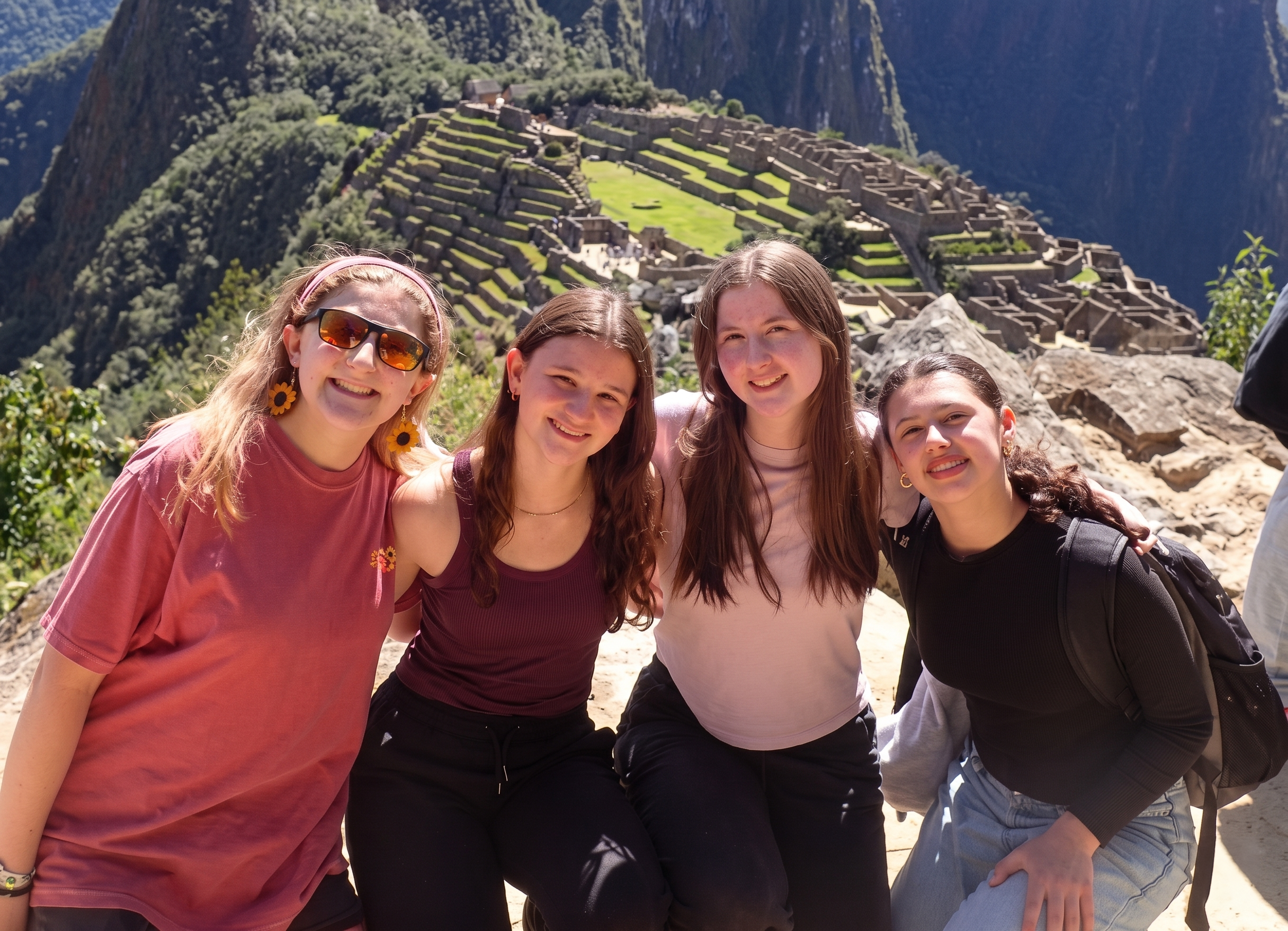Four young women pose and smile together in front of the ancient ruins of Machu Picchu in Peru, with sunlight highlighting the stone terraces and green mountain scenery in the background.