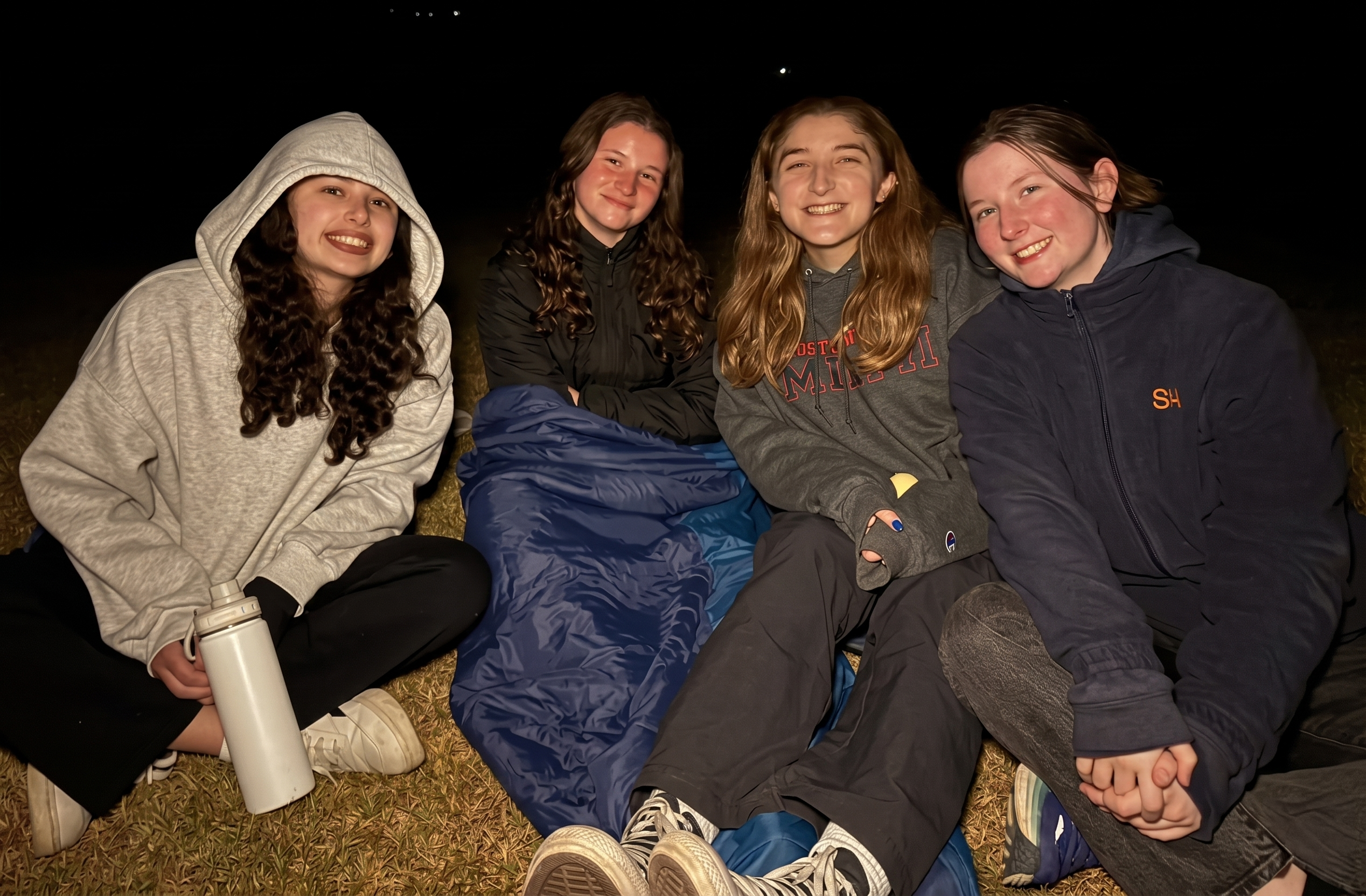 Four teenage girls sit together on grass at night, smiling at the camera. One wears a hoodie and holds a water bottle, another is wrapped in a sleeping bag, and all are dressed warmly. The background is dark.