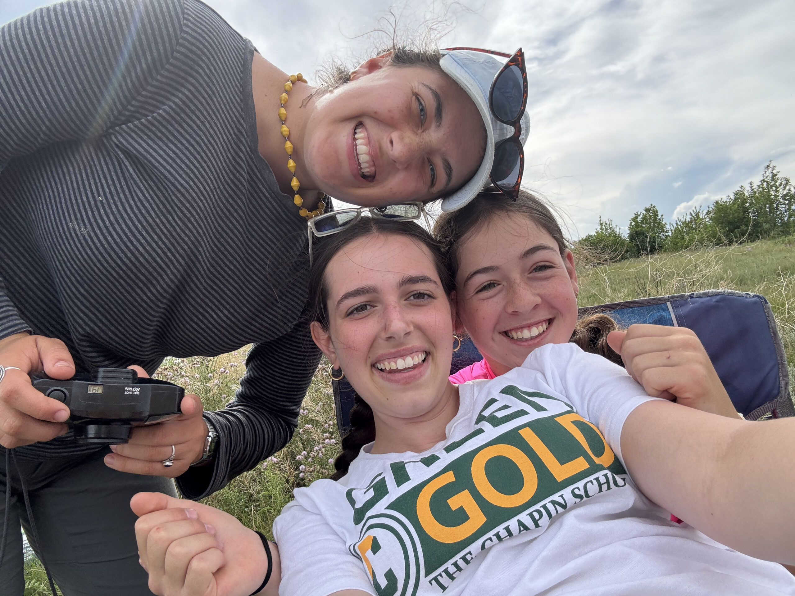 Three smiling young women, including VISIONS alumni, pose for a cheerful outdoor selfie. One wears a Green Gold T-shirt, another holds a camera. They appear happy and relaxed on a sunny day with grass and sky in the background.