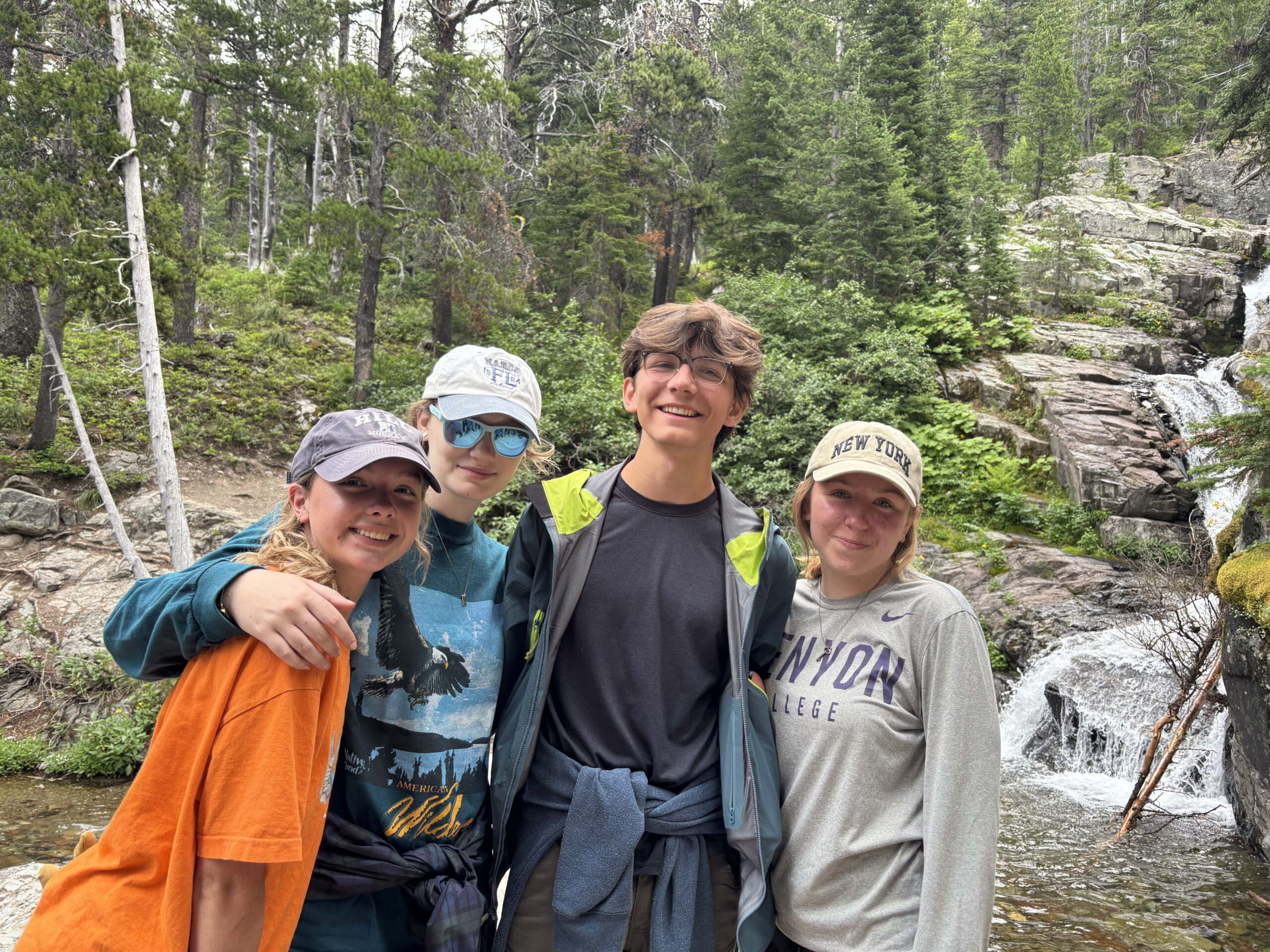 Four smiling teens stand close together in front of a small waterfall and lush green forest, wearing casual outdoor clothes and baseball caps. It appears to be a hiking trip in nature.