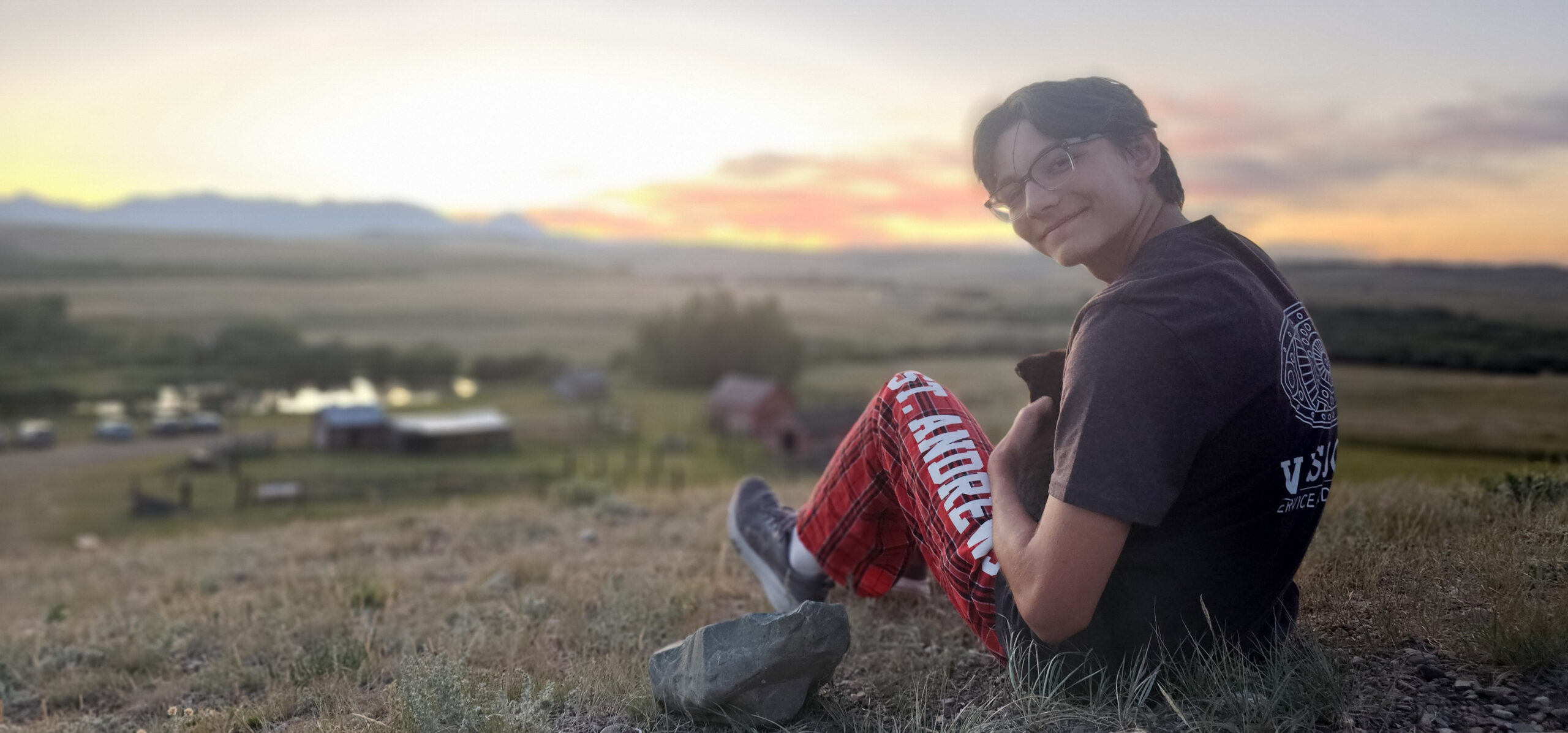 A person in glasses and red plaid pants sits on a grassy hill at sunset, looking back at the camera. The background shows open fields, a pond, scattered buildings, and distant mountains under a colorful sky.