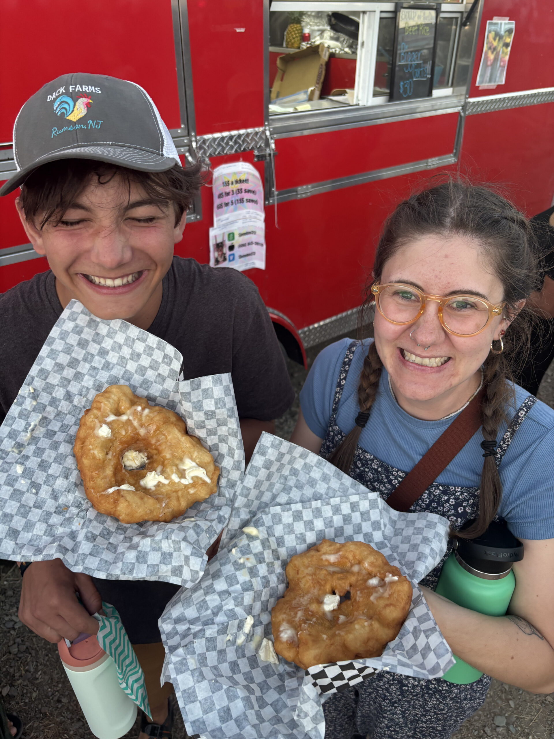 Two smiling people hold up fried dough covered in butter on checkered paper in front of a bright red food truck. One wears a gray cap; the other wears glasses and pigtails. Both hold water bottles.