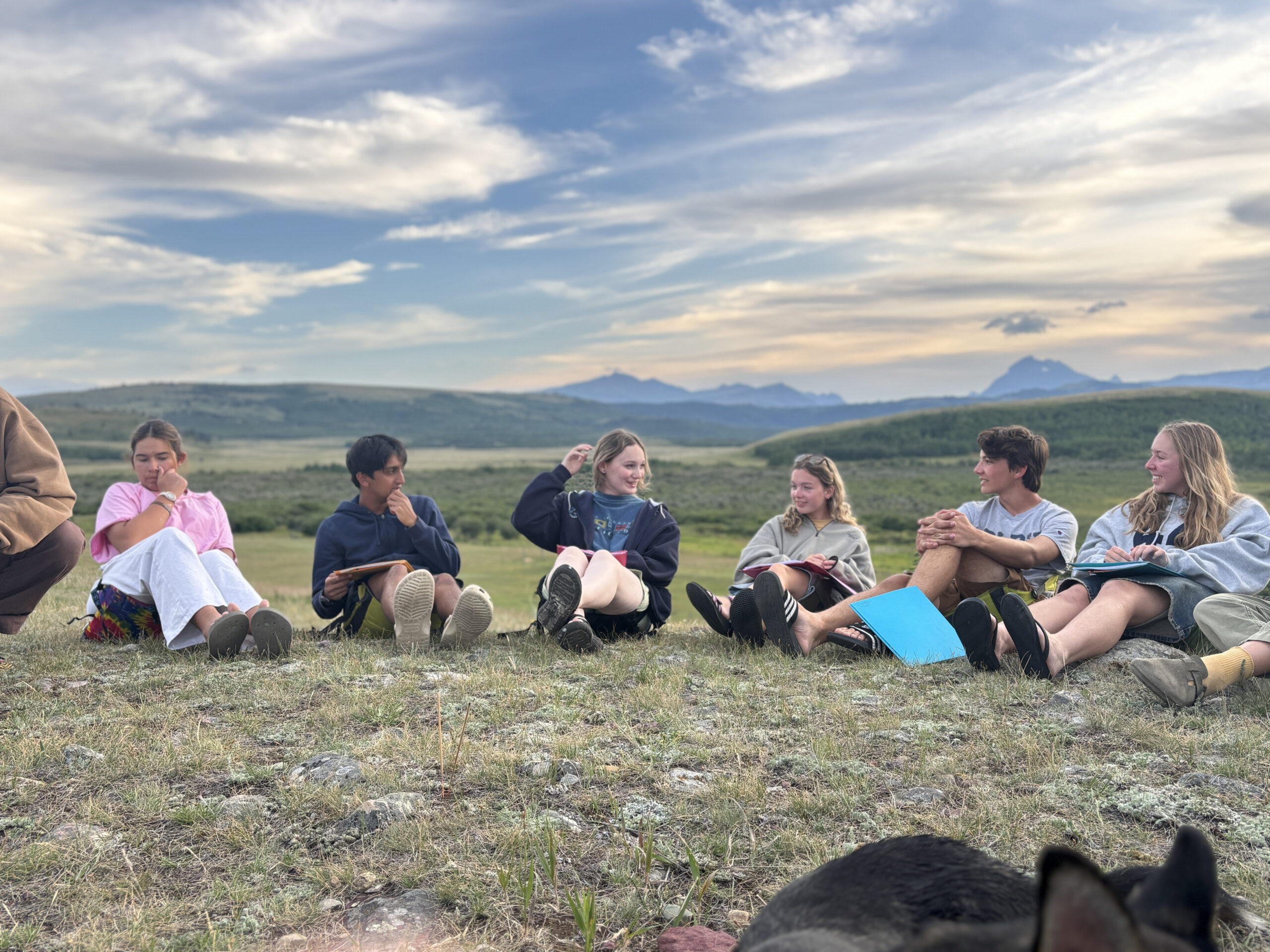 A group of six young people sits in a circle on grass with notebooks, talking and smiling. Rolling hills and mountains are visible in the background under a cloudy sky. A dog’s ears appear at the bottom of the image.