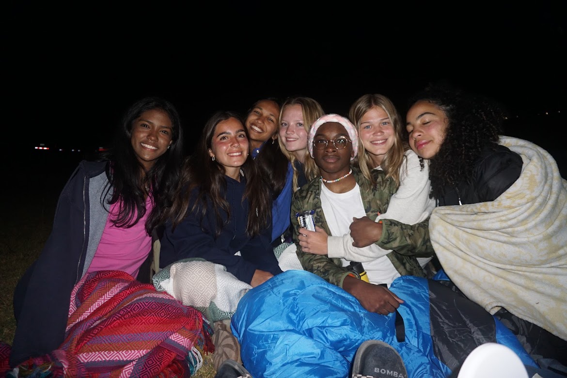 Seven friends sit closely together outdoors at night, smiling and wrapped in cozy blankets. They appear relaxed and happy, illuminated by a camera flash against a dark background.