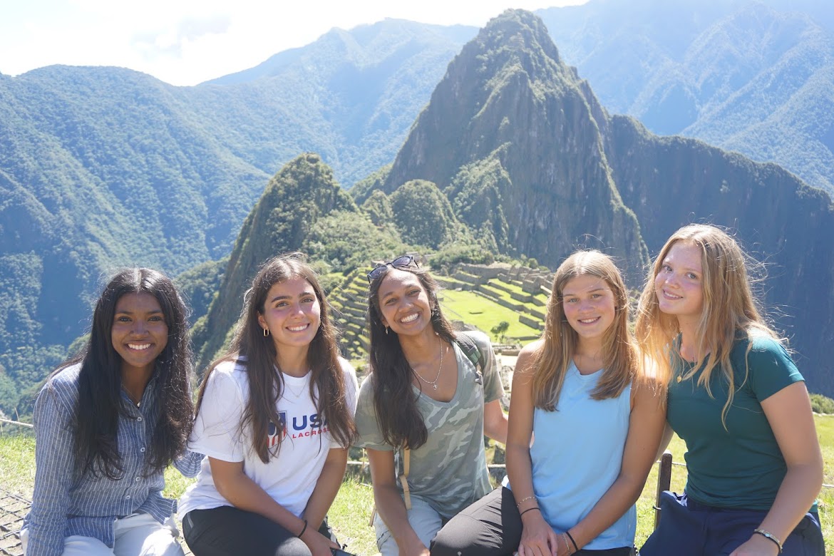 Five young women smile while sitting together in front of the ancient ruins of Machu Picchu, with green mountains and blue sky in the background.