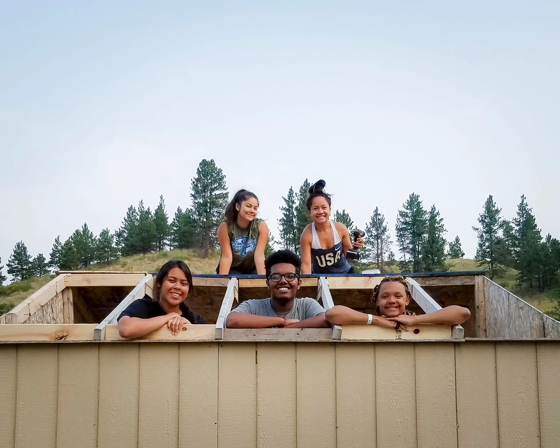 Group of teens poke their heads our of the top of a structure before building the roof on community service trips to the Northern Cheyenne