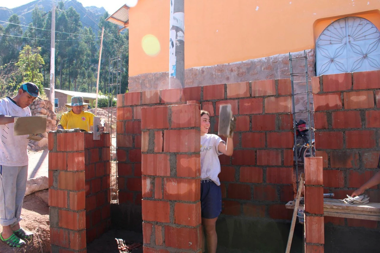 Teens and local builders laying bricks and applying mortar at an outdoor construction site in Peru, with mountains and trees in the background - VISIONS Summer Program